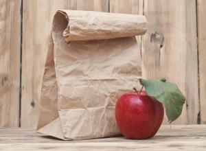 Paper bag on a desk with an apple beside it.