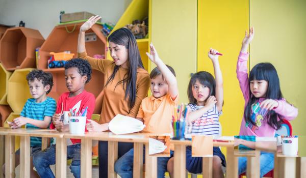 Children in a row, sitting at their desks, with early years provider. Some are raising their hands.